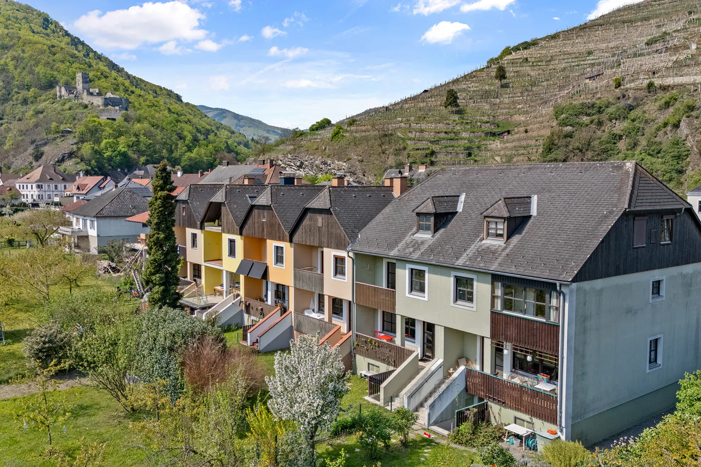 Row of colorful townhouses with dark roofs, set against a backdrop of green hills and vineyards under a blue sky.