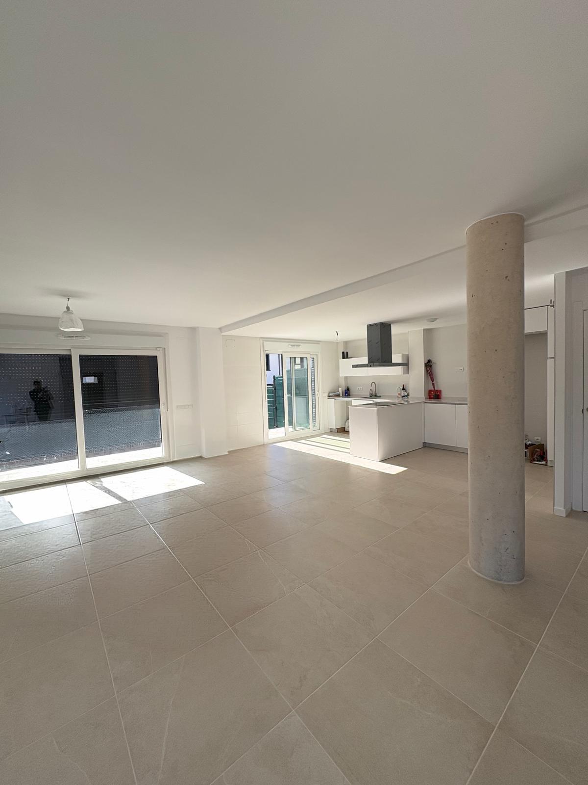 Bright, open-plan living space with beige tile flooring, white walls, and a modern kitchen with island. Large windows let in sunlight.