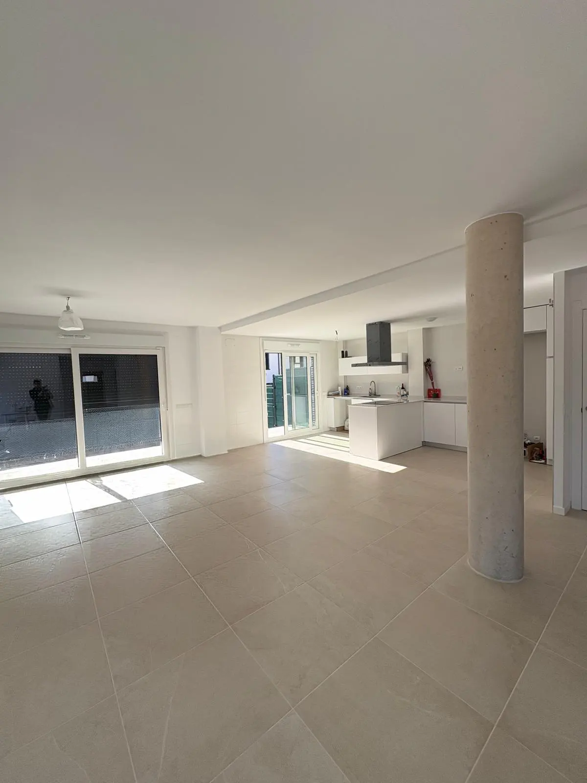 Bright, open-plan living space with beige tile flooring, white walls, and a modern kitchen with island. Large windows let in sunlight.