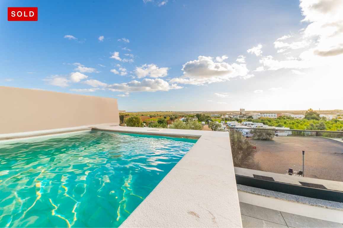 Rooftop infinity pool with turquoise water overlooking a sunny landscape with trees and a parking area under a blue sky. [AI-generated text]