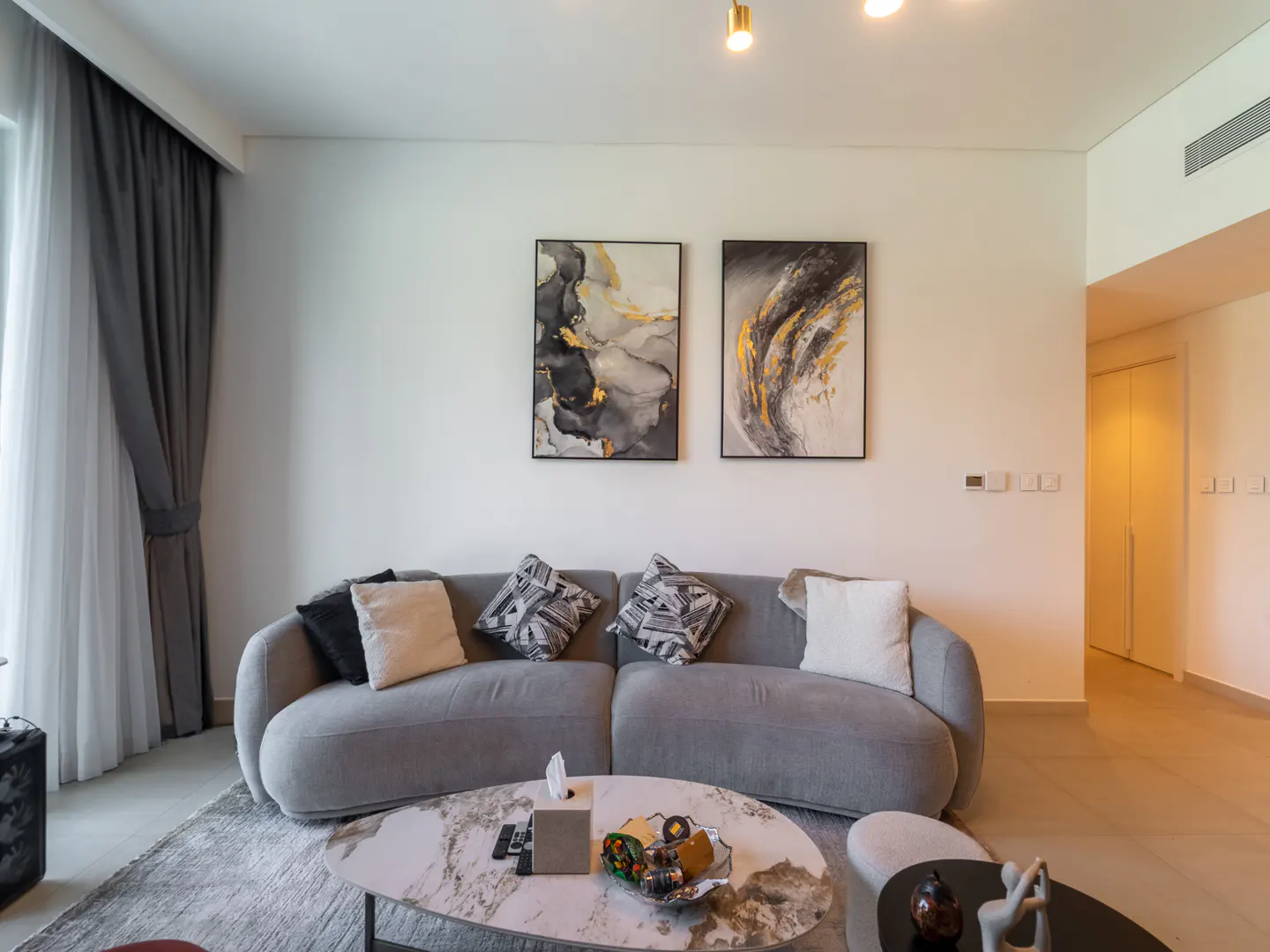 Living room with a gray curved sofa, marble coffee table, and abstract art on the white wall. Gray curtains frame the window.