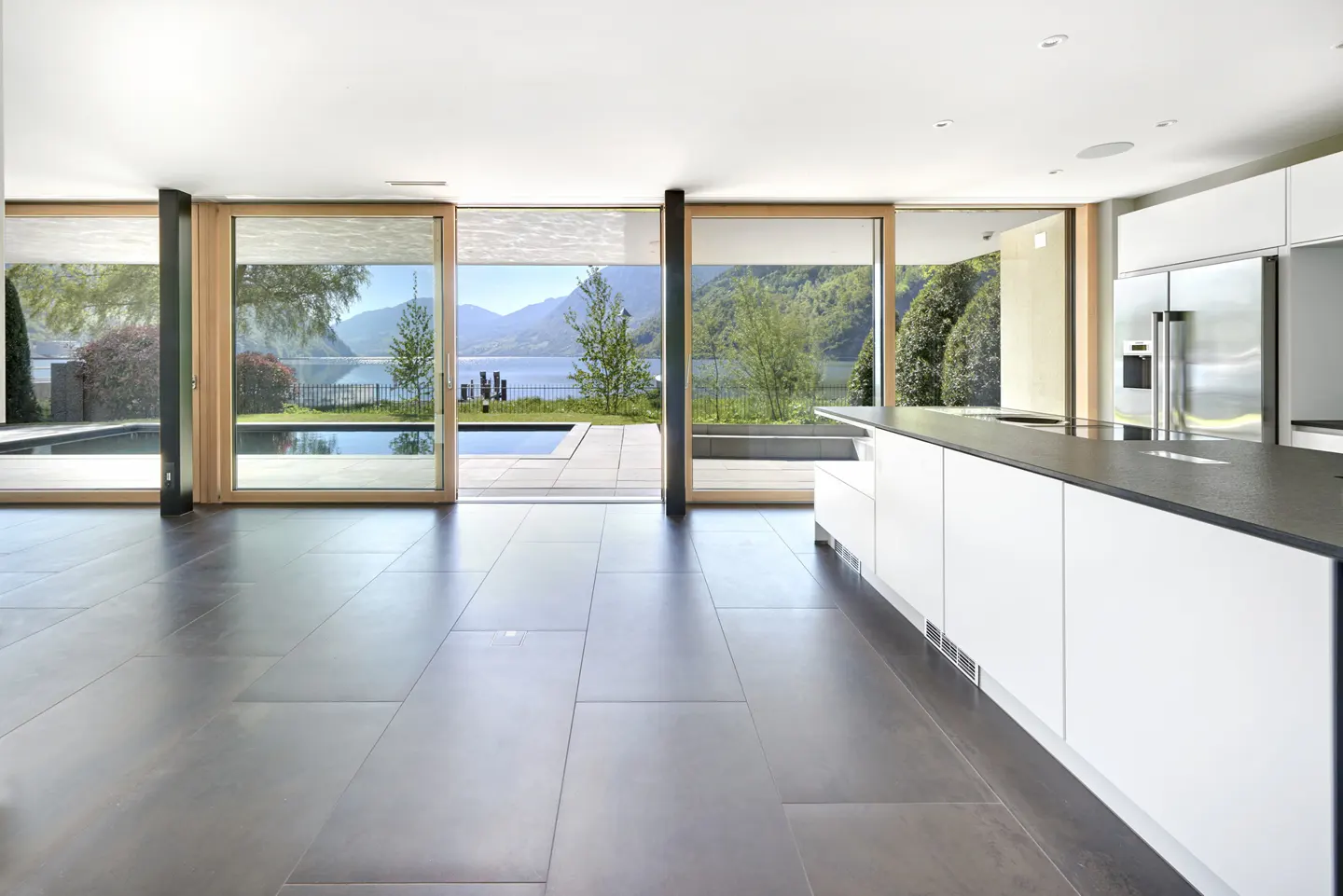 Modern kitchen with dark tile floors and white cabinets. Large sliding doors open to a pool and lake view.