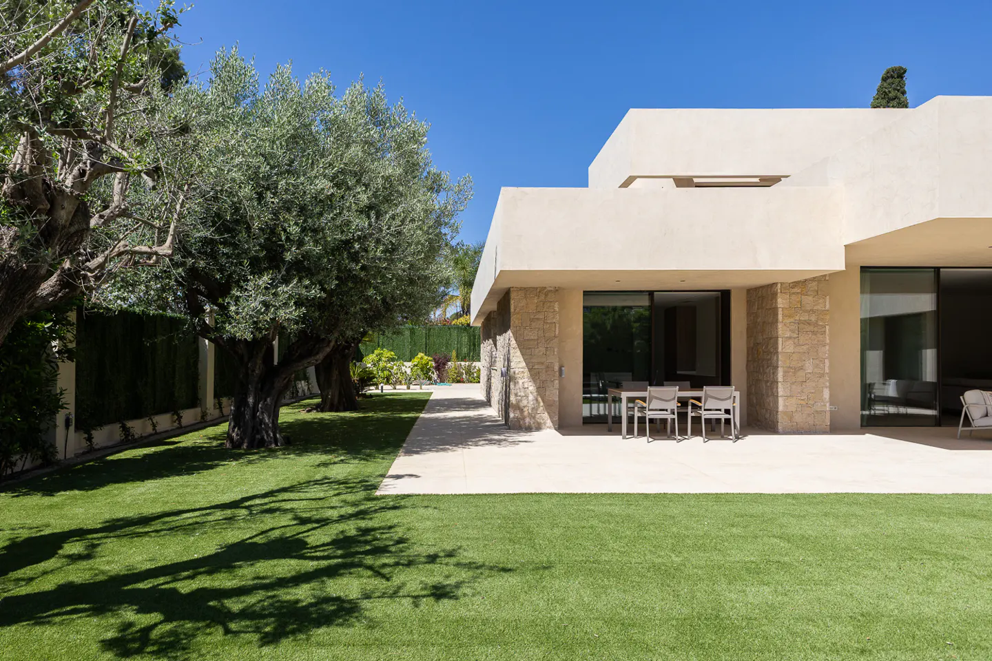 Modern beige house with a patio, table, and green lawn under a clear blue sky. Olive trees cast shadows on the grass.