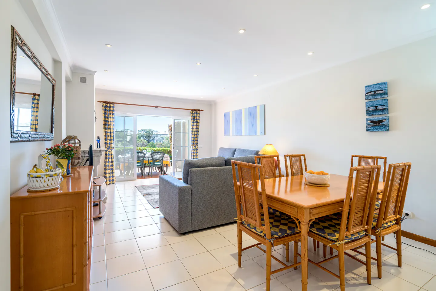 Bright living and dining area with white walls, tile floors, and a gray sofa. A wooden table with six chairs sits near a balcony with outdoor seating.