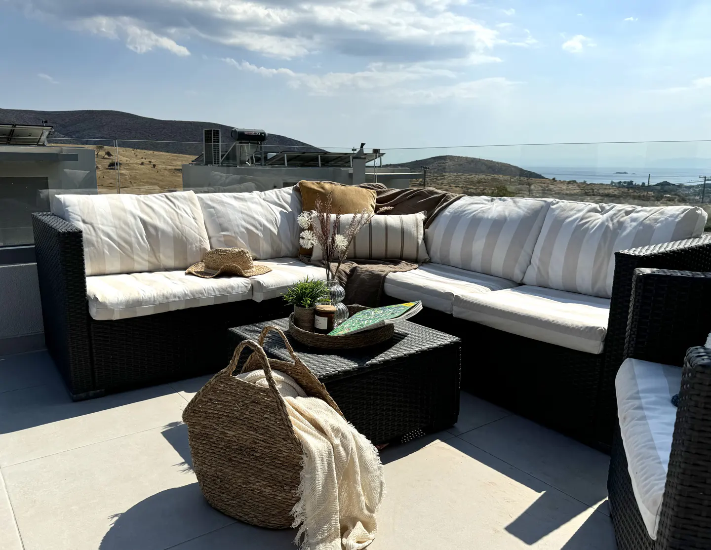 Outdoor patio with wicker furniture, white cushions, and a woven basket with a blanket. A table holds plants and a book, with a scenic view in the background.