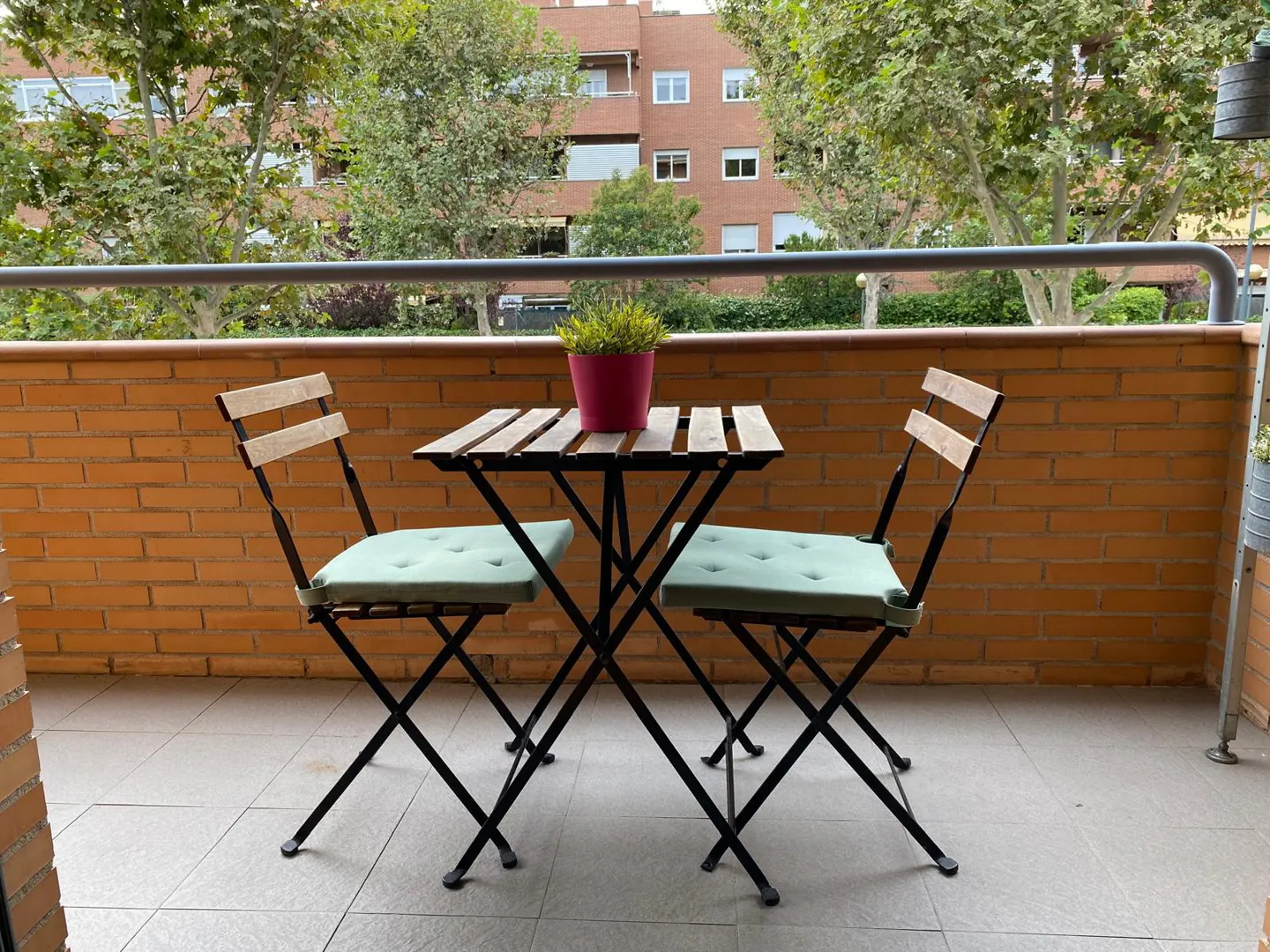 A small wooden bistro table with a pink potted plant and two chairs with light green cushions on a brick-walled balcony.