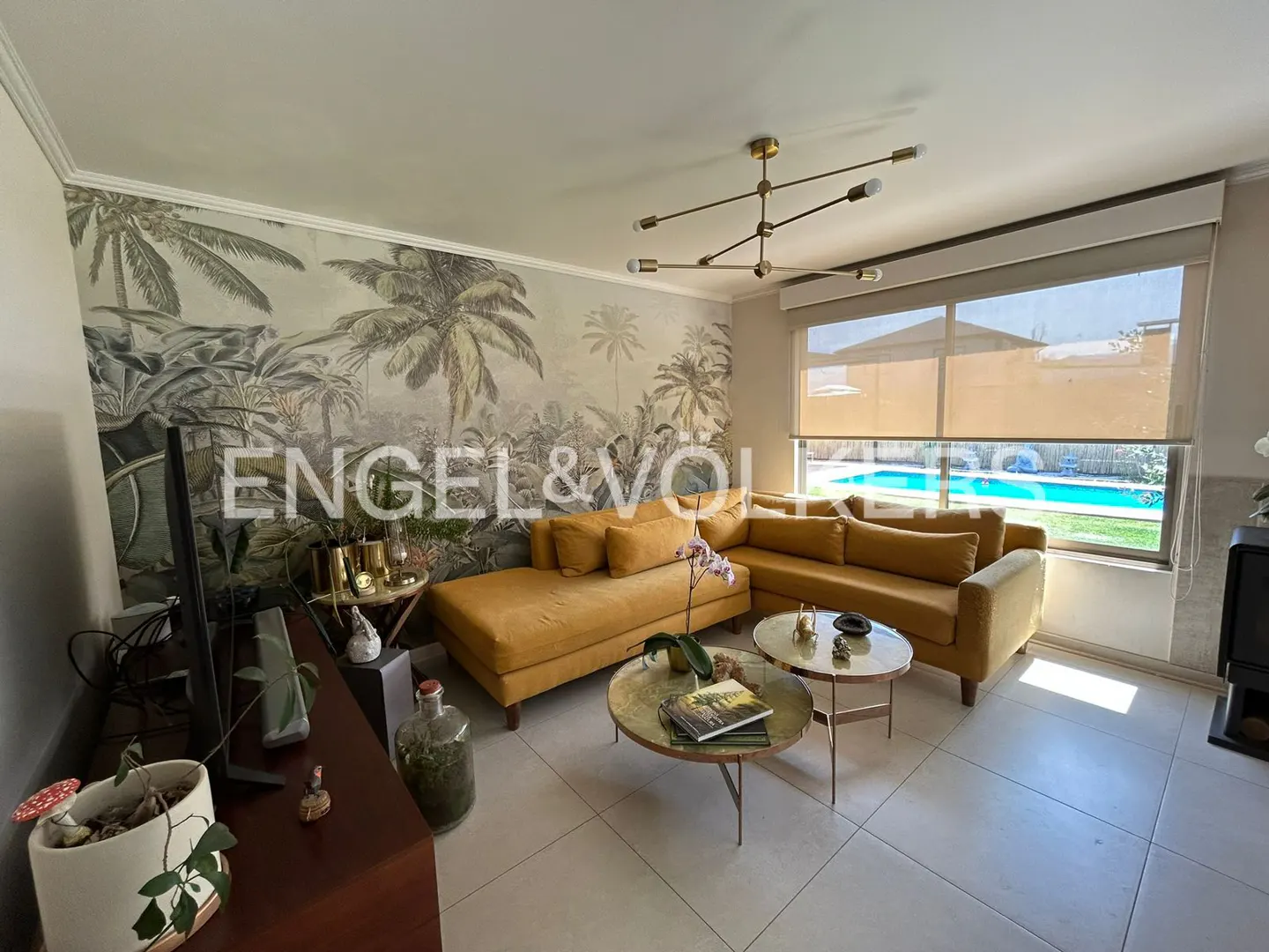 Living room with mustard sectional sofa, round tables, and tropical wallpaper. A pool is visible through the window.