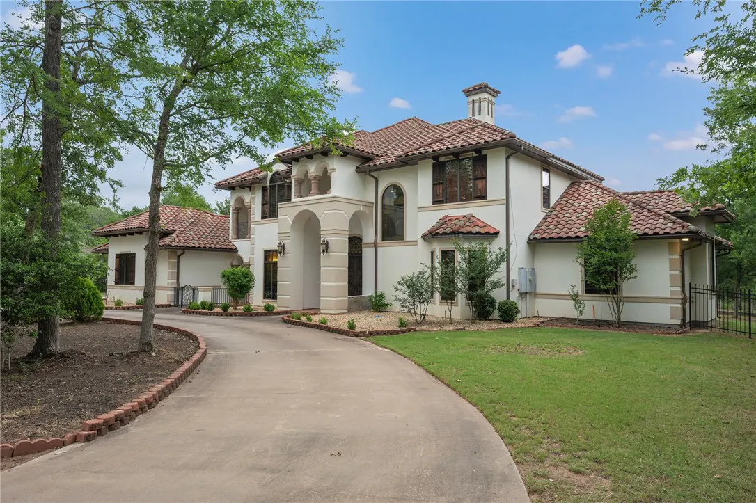 Exterior view of a two-story, white stucco house with a red tile roof and arched entryway. A curved driveway leads to the home, surrounded by green trees and lawn.