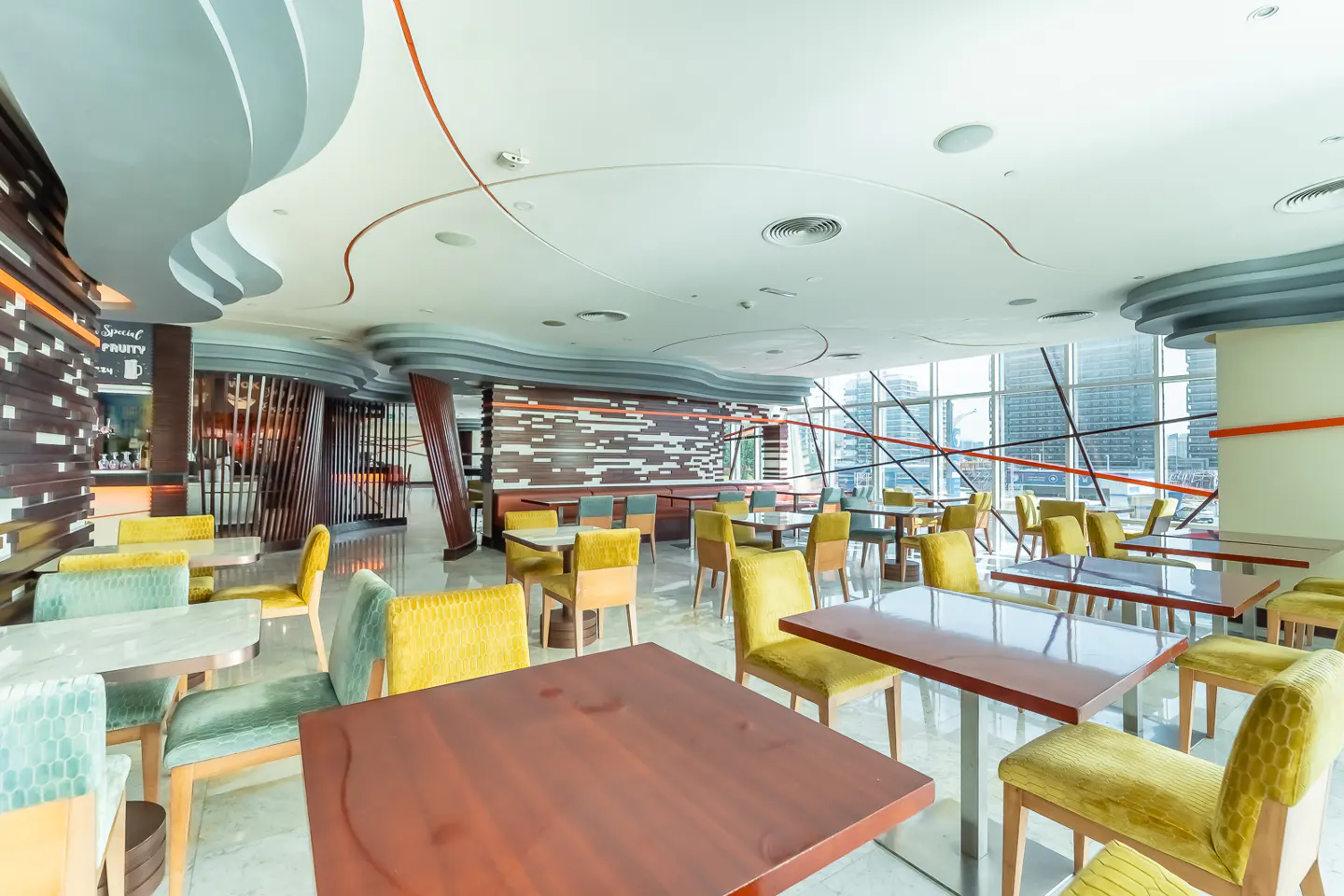 Bright restaurant interior with wood tables, yellow and green chairs, and a city view through large windows.