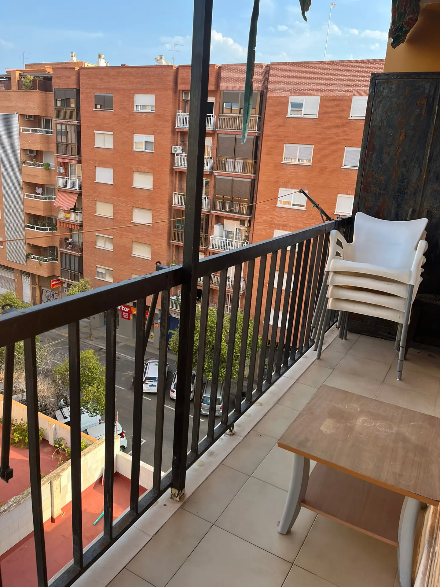 Balcony view with black railings, stacked white chairs, and a wooden table. Brick buildings and a street are visible in the background.