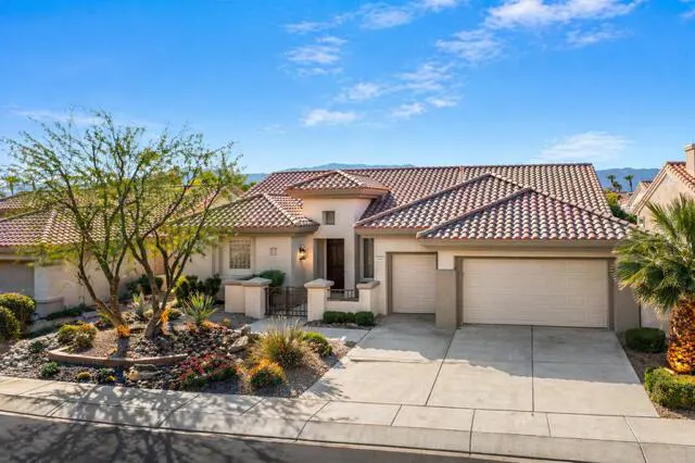 Beige single-story house with a red tile roof, two-car garage, and desert landscaping under a blue sky.