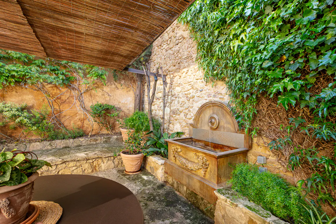 Outdoor patio with stone walls covered in ivy, a stone fountain, potted plants, and a brown table under a bamboo shade.