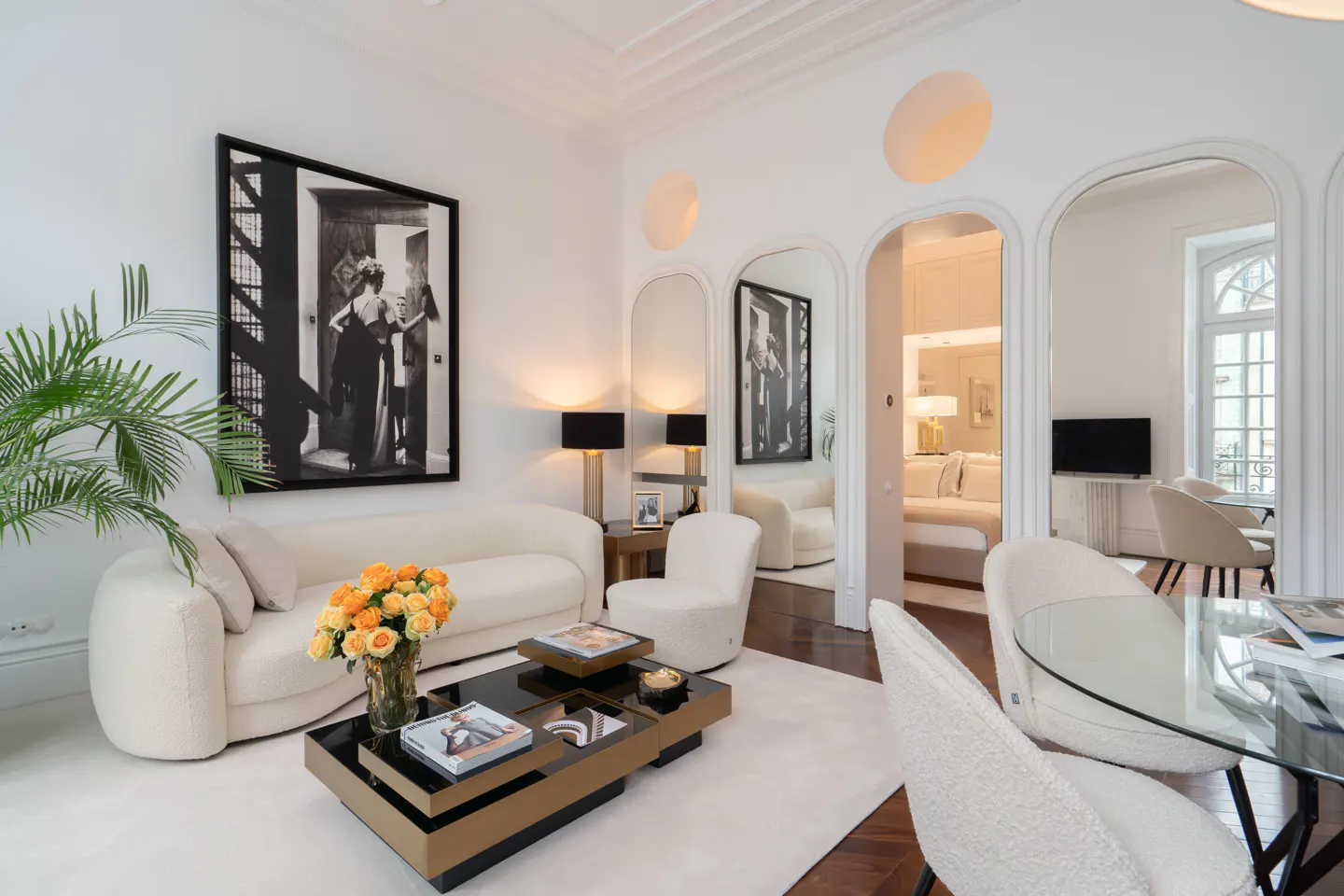 Bright living room with white walls, sofa, and chairs. A black and gold coffee table sits on a white rug. Arched doorways lead to other rooms.