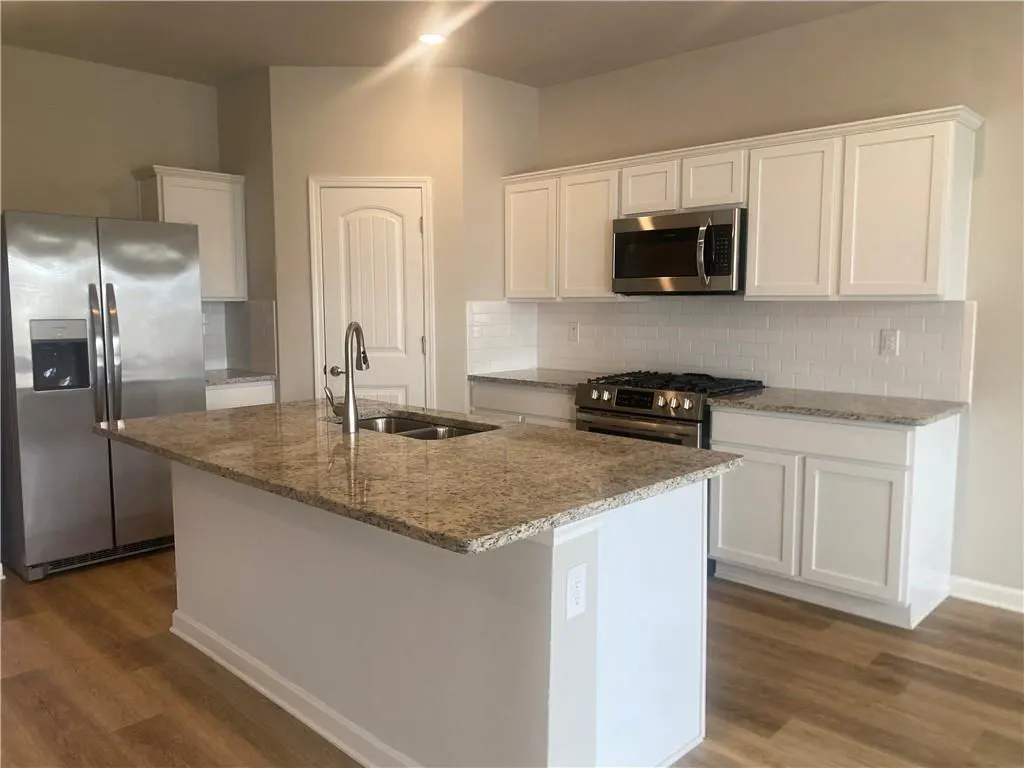 Bright kitchen with white cabinets, stainless steel appliances, and a granite island with a sink. Wood-look flooring.