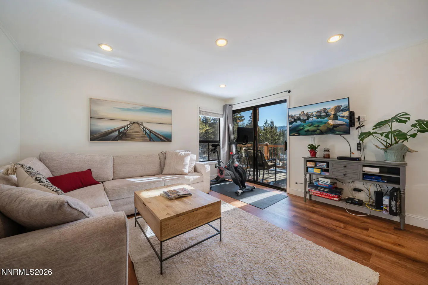 Living room with a beige sectional sofa, wood coffee table, TV, exercise bike, and sliding glass doors to a deck.