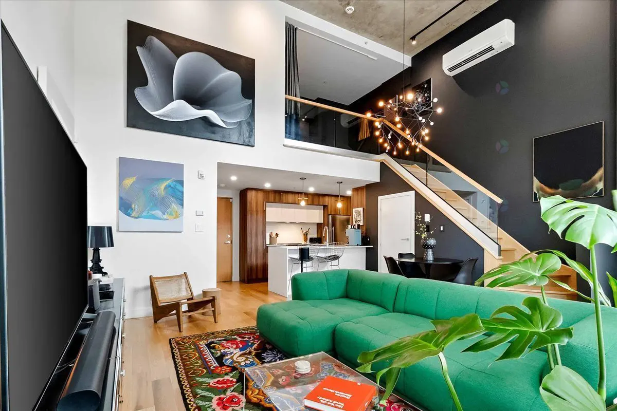 Bright living room with a green sofa, wood floors, and a black accent wall with a modern chandelier. Stairs lead to a loft area.