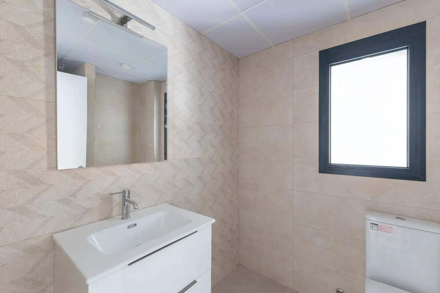 Modern bathroom with beige chevron-patterned tiled walls, a white vanity sink, a mirror, and a window with a black frame.