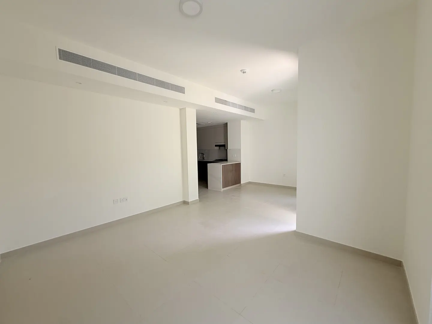 An empty, bright room with white walls and light gray tile floors. A kitchen area is visible in the background.