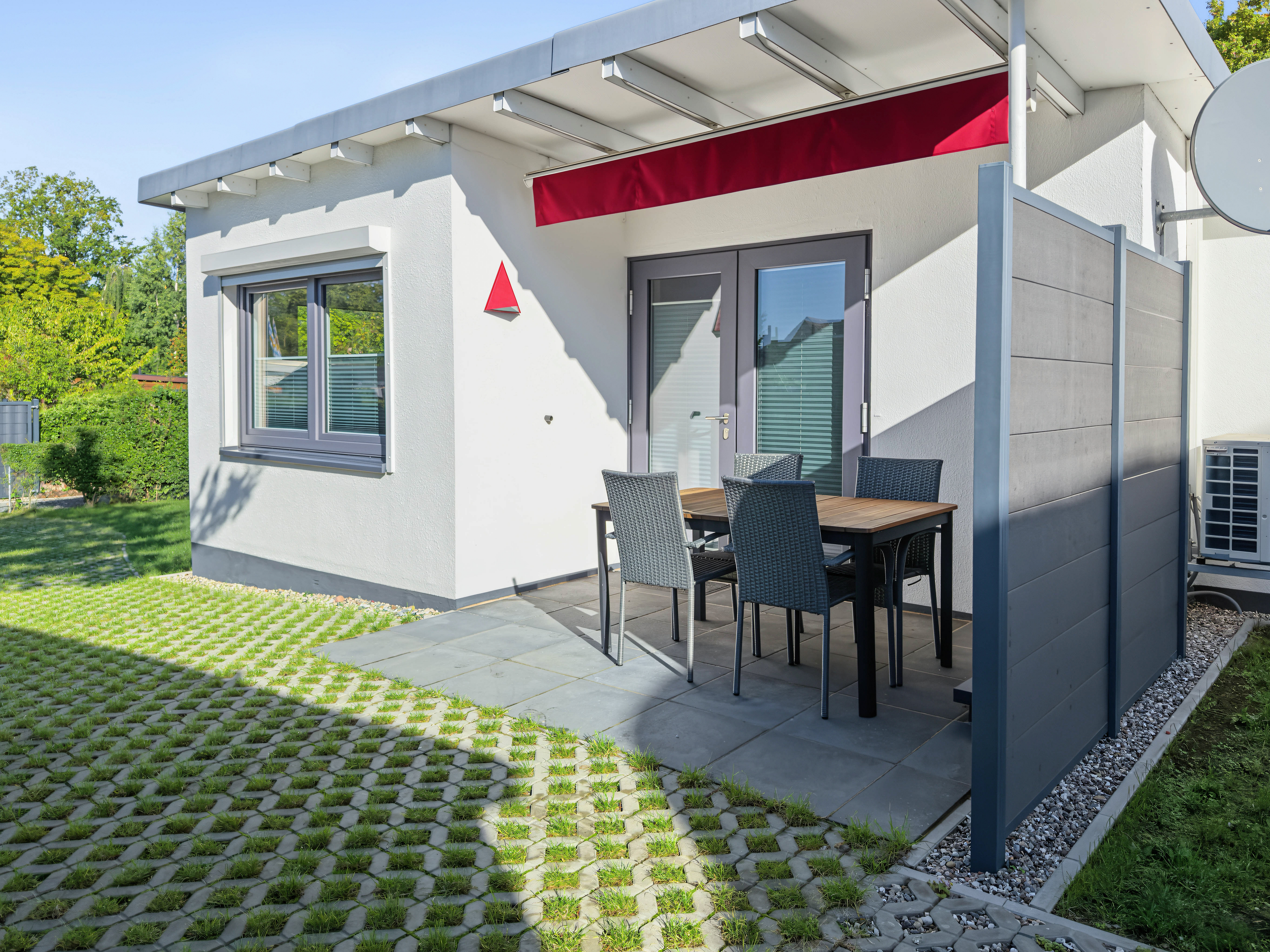Modern white house exterior with a patio, dining set, red awning, and a green lawn with paved grid stones. [AI-generated text]
