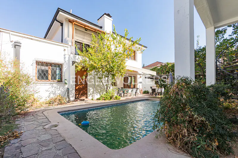 Exterior view of a two-story white house with a pool, stone patio, and green foliage. A blue ball floats in the pool.