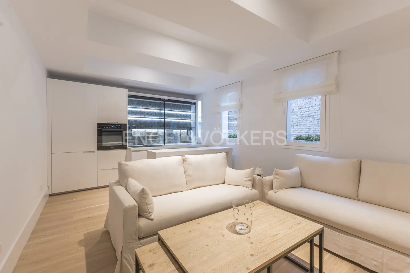 Bright, modern living room with white sofas, wood coffee table, and kitchen area with white cabinets. Windows with white blinds let in natural light.