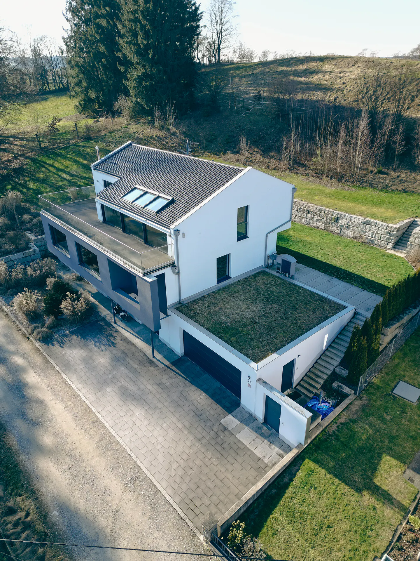Aerial view of a modern white house with a green roof, dark gray trim, and a paved driveway. Trees and a grassy hill are in the background.
