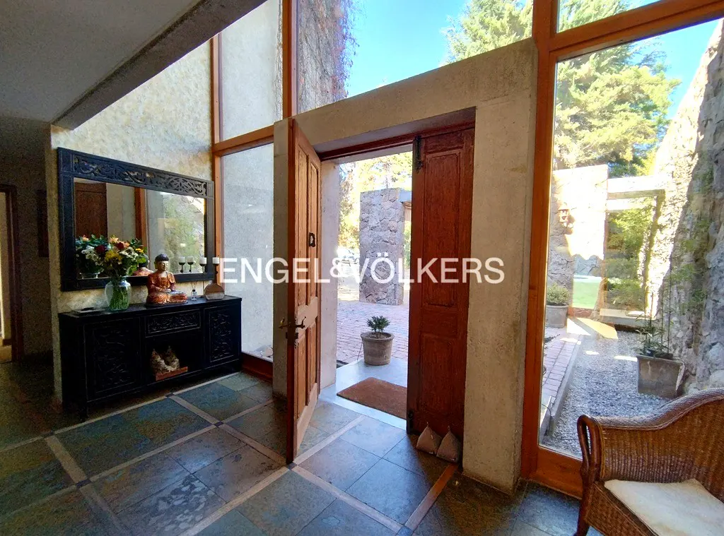 Open wooden front door of a home with stone walls and large windows. A black console table with a mirror and Buddha statue is visible.