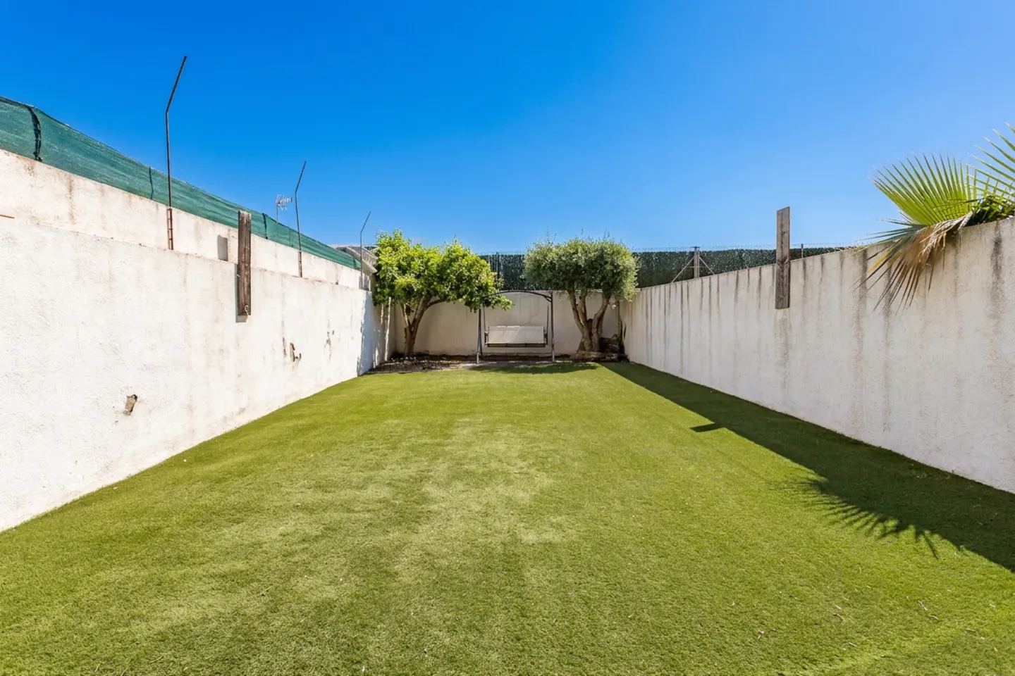 A backyard with green grass, white walls, and a swing set between two trees under a clear blue sky.