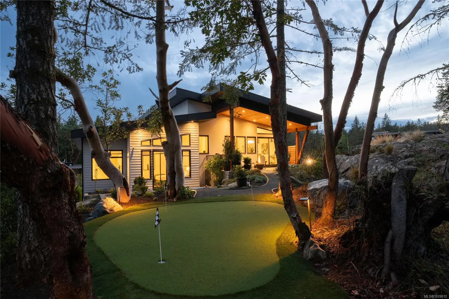 Exterior view of a modern home with a putting green, seen through trees at dusk.