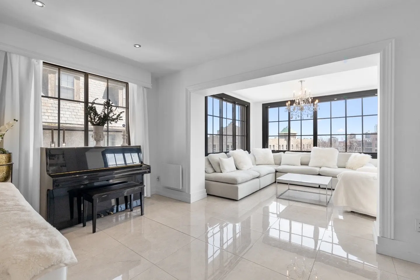 Bright, open-concept living room with a black piano, white sofa, and large windows with city views.