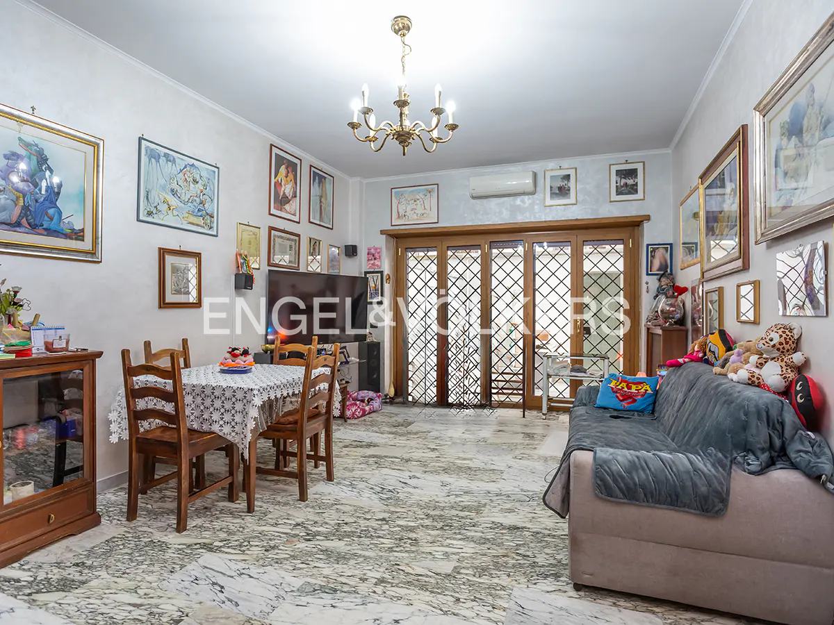 Living room with marble floors, a dining table with four chairs, and a sofa with pillows and stuffed animals. Walls are decorated with framed art.