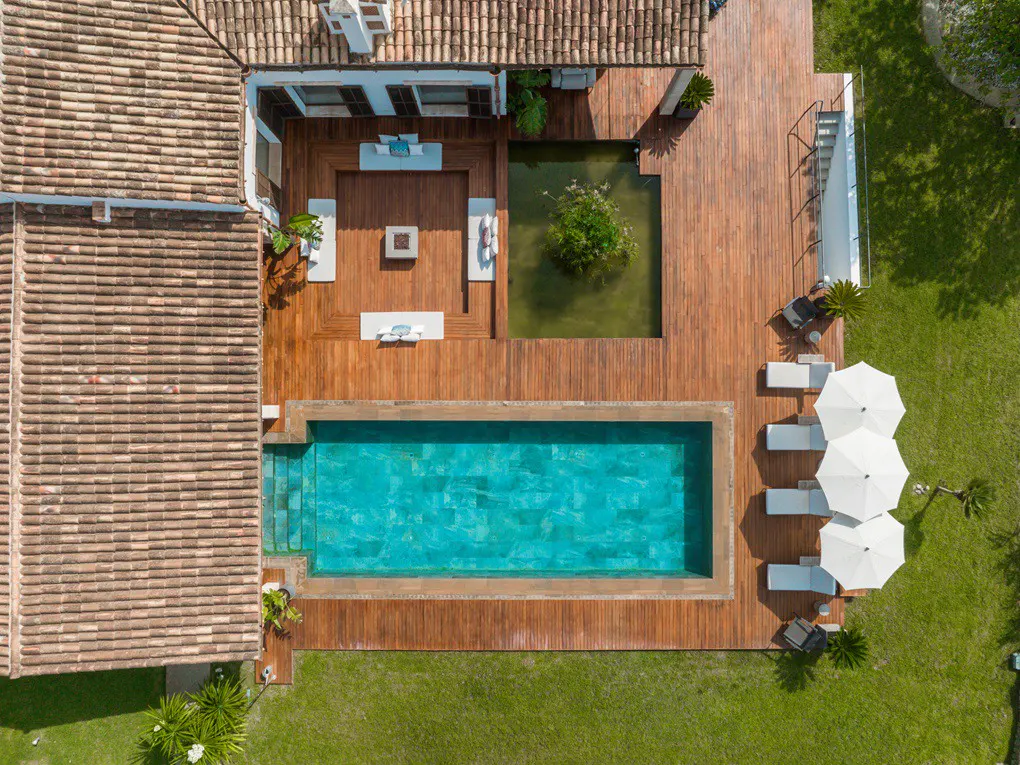 Aerial view of a luxury home with a pool, pond, and wooden deck. White lounge chairs and umbrellas sit on the green lawn.
