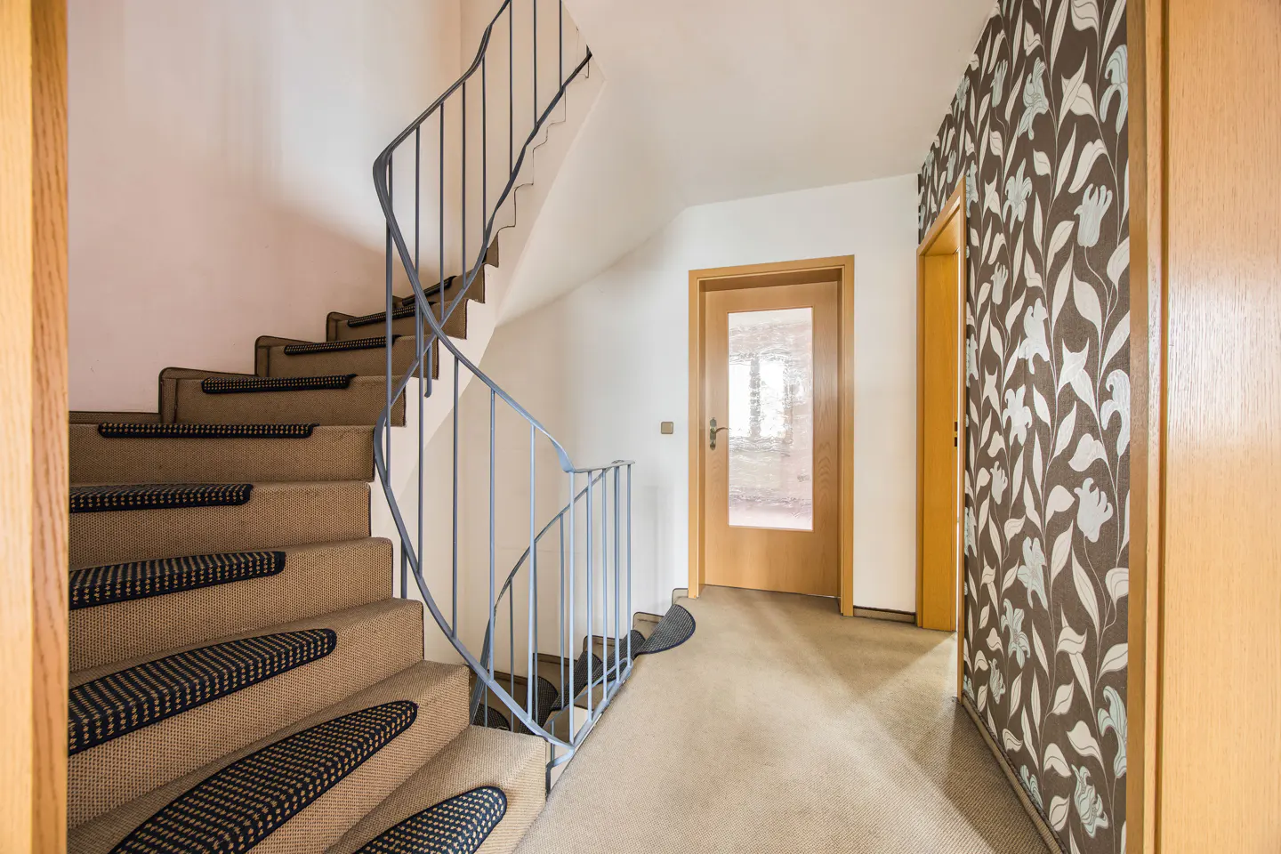 Staircase with beige carpet and dark patterned treads, featuring a blue metal railing in a bright, modern hallway.