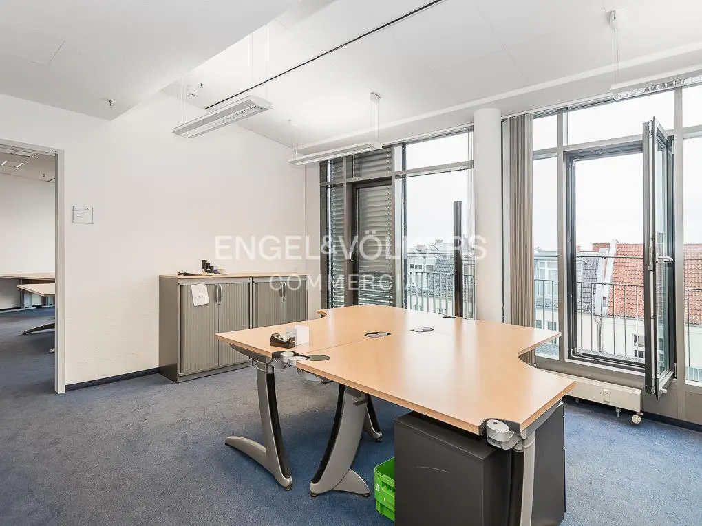 Bright office space with blue carpet, a large desk, and a storage cabinet. Windows offer a city view. Engel & Völkers logo visible.