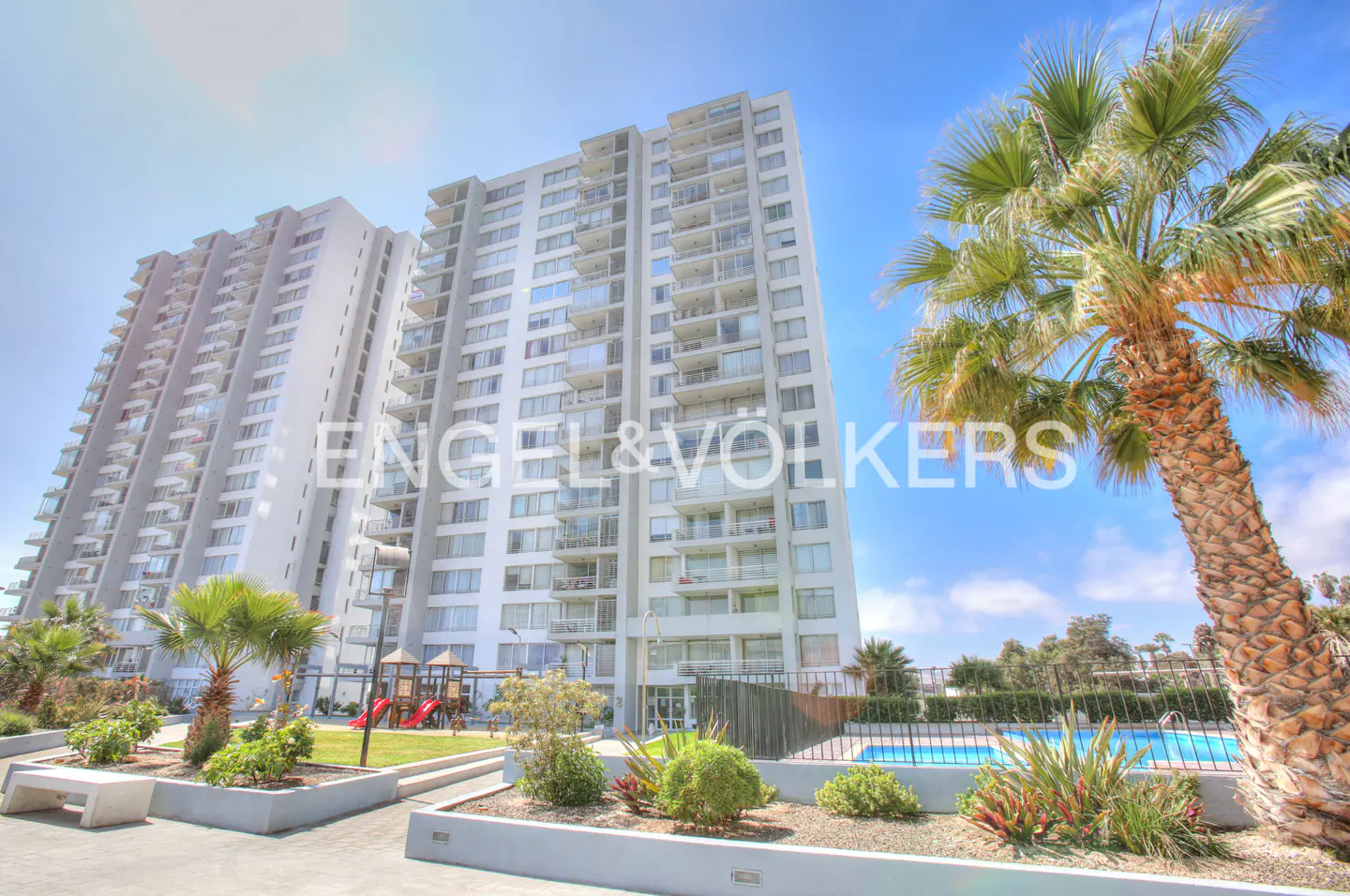 Exterior view of two tall, white apartment buildings with balconies, a pool, and palm trees under a blue sky.