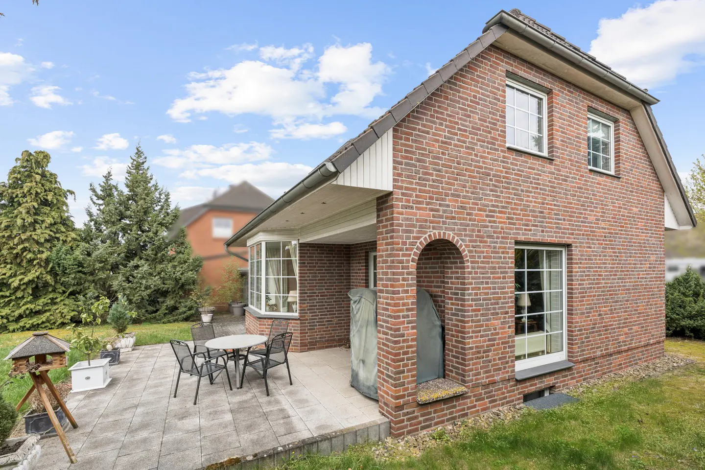 Brick house with a patio featuring a table and chairs, surrounded by green grass and trees under a cloudy sky.