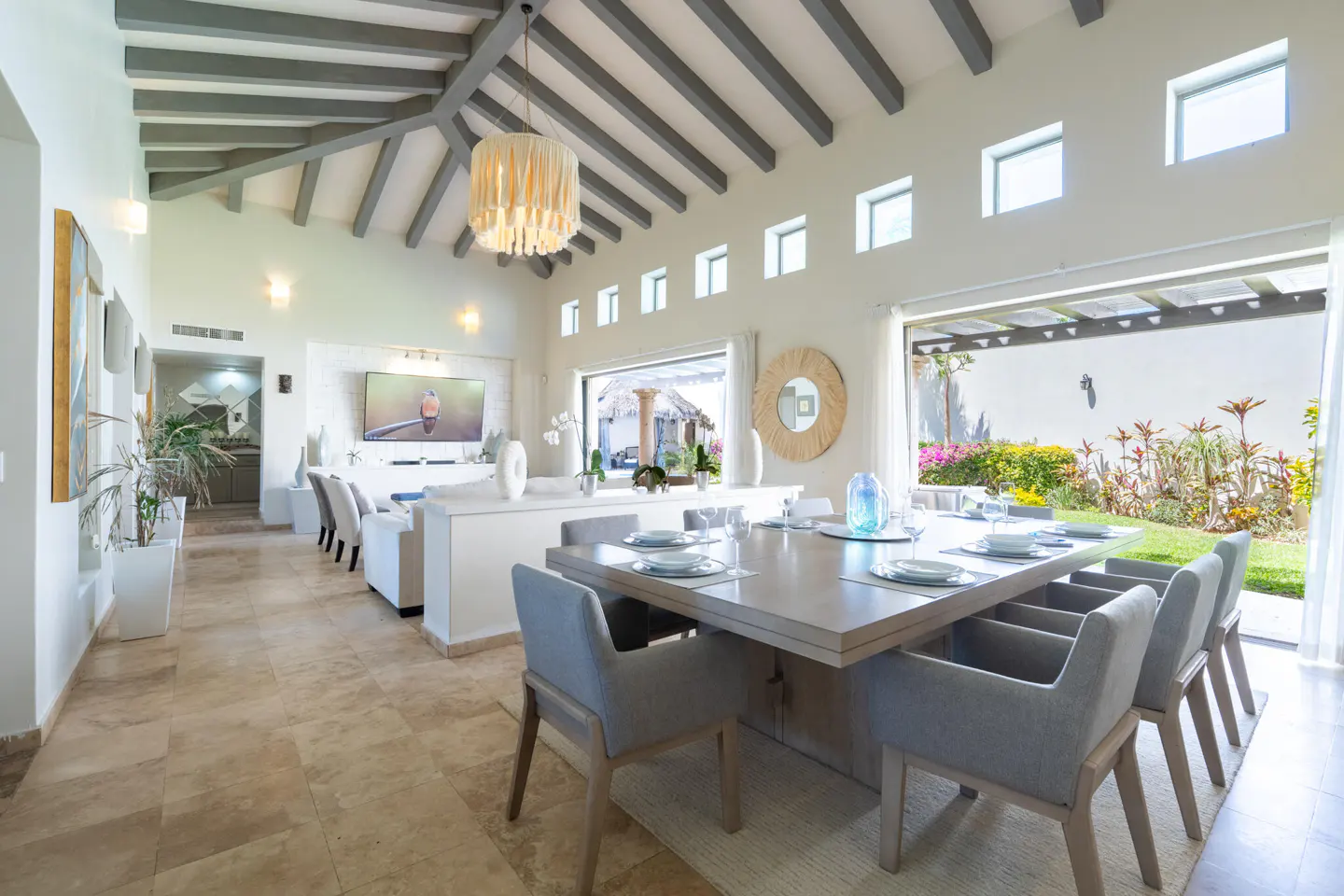 Bright, open-plan living and dining area with a large table set for a meal. Outside, a lush garden is visible through sliding glass doors.