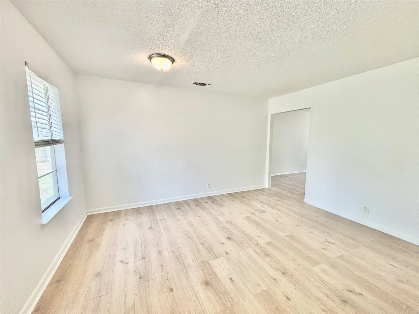 Bright, empty room with light wood floors, white walls, and a window with blinds. A doorway leads to another room.