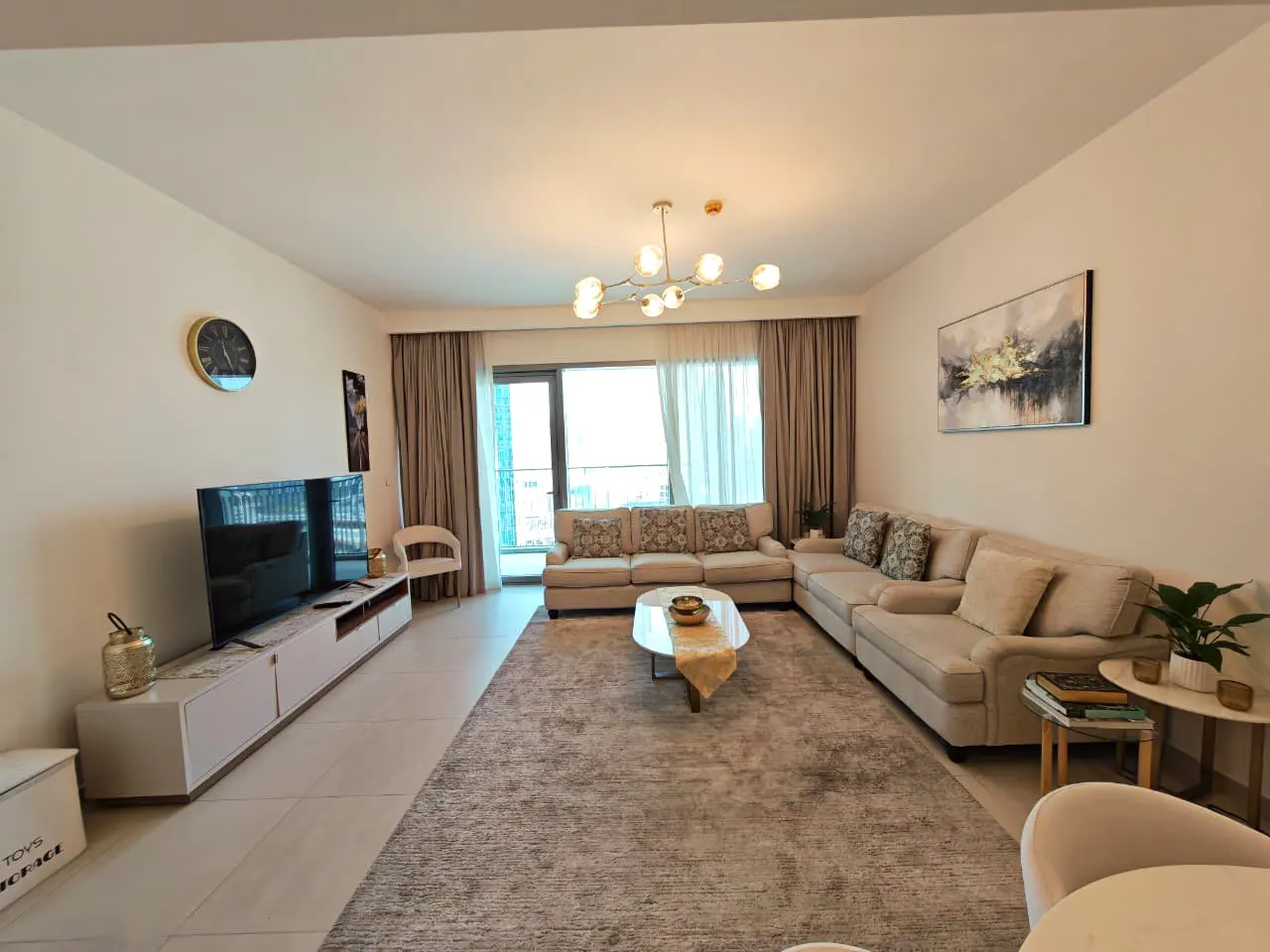 Living room with a large beige sectional sofa, a gray rug, and a TV on a white console. A modern chandelier hangs from the ceiling.
