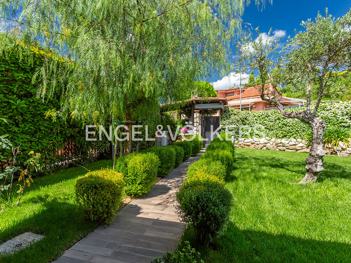Landscaped garden with a stone path lined by green bushes leading to a gate, with a house visible in the background.