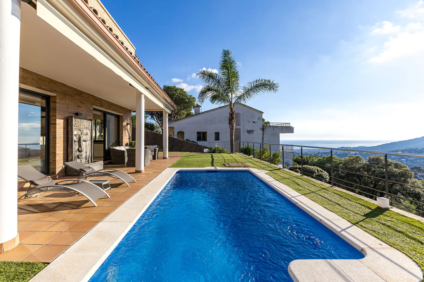 A bright blue pool sits beside a patio with lounge chairs, under a covered porch of a brick house, with a palm tree and ocean view.