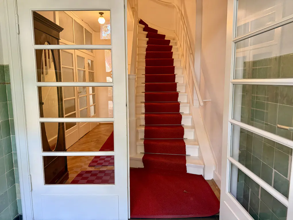 Foyer view with white staircase covered in red carpet. A glass door shows a glimpse of a checkered floor and a dark wood cabinet.