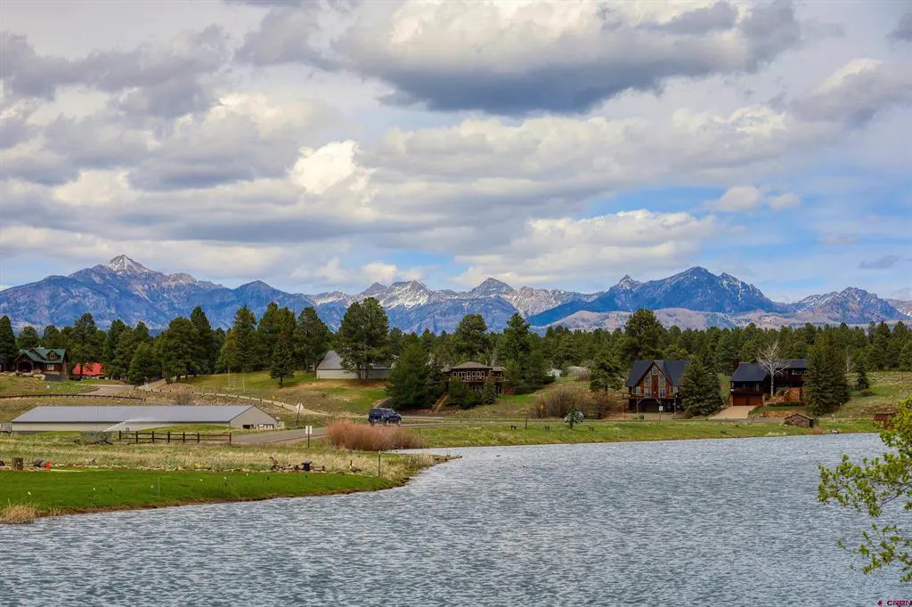 Scenic view of a lake with houses, trees, and snow-capped mountains under a cloudy sky.