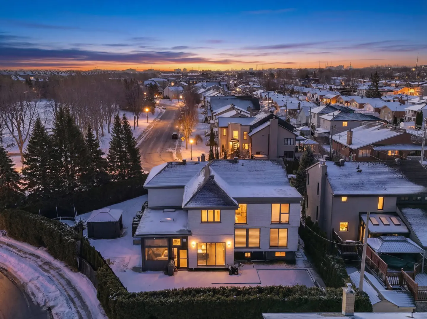 Aerial view of a modern, two-story white house with snow-covered roof and yard at dusk. Streetlights illuminate the snowy neighborhood.