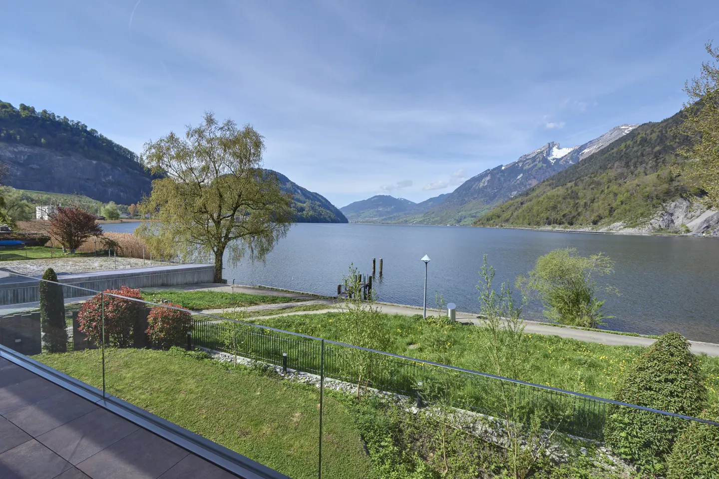 Scenic view of a lake surrounded by mountains, seen from a balcony with a glass railing and green lawn.