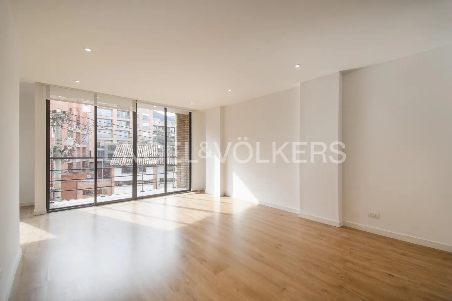 Bright, empty room with light wood floors, white walls, and a large window showing a city view. Engel & Völkers logo on the wall.