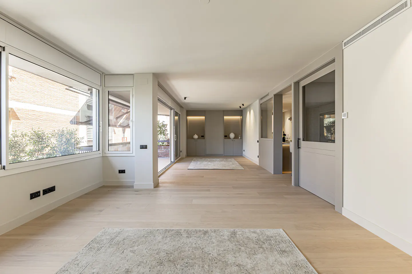 Bright, empty room with light wood floors, large windows, and sliding glass doors to a balcony. Gray built-in cabinets and doors add subtle color.