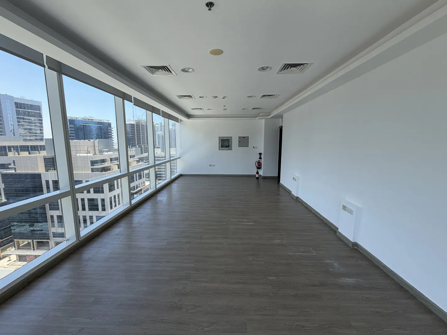Empty office space with wood-look flooring, white walls, and floor-to-ceiling windows overlooking city buildings.