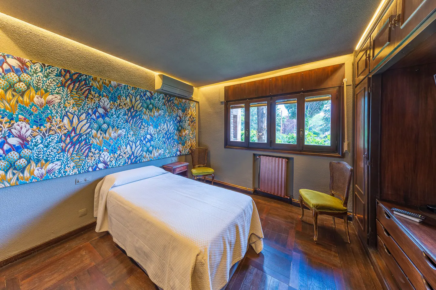 Bedroom with white bed, floral wall art, and hardwood floors. Two chairs with green cushions flank a window. A wooden wardrobe stands to the right.