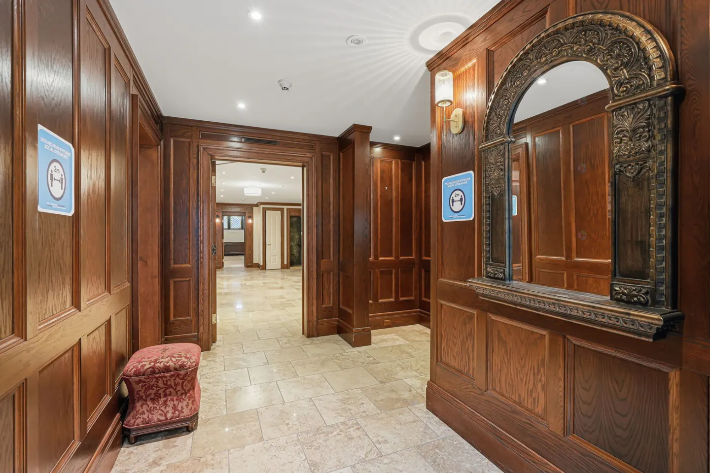 Hallway with dark wood paneling, beige tile floor, and ornate mirror. A red patterned stool sits near a doorway. Social distancing signs are posted.